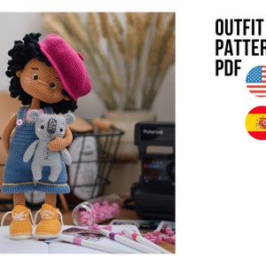 Puede incluir: Muñeca de crochet con piel morena, cabello negro y boina rosa que sostiene un koala gris. La muñeca lleva un vestido de peto vaquero azul con costuras amarillas. La muñeca está sentada en una mesa con una cámara Polaroid y cuentas rosas de fondo.  OUTFIT PATTERN PDF está escrito en texto negro sobre fondo blanco. Las banderas estadounidense y española se muestran en la imagen.