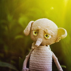 Puede incluir: Un juguete de peluche gris tejido a crochet con grandes ojos verdes y una nariz larga. El juguete est&aacute; en un fondo verde y amarillo.