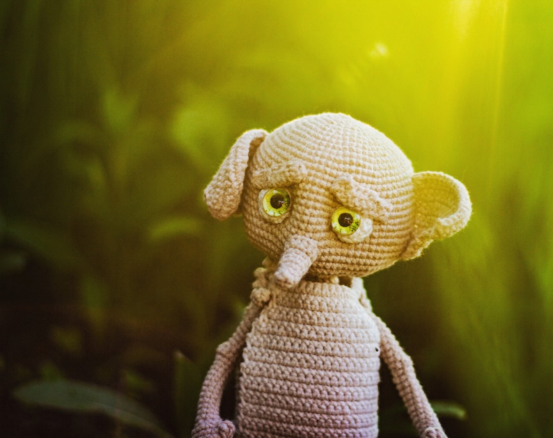 Puede incluir: Un juguete de peluche gris tejido a crochet con grandes ojos verdes y una nariz larga. El juguete est&aacute; en un fondo verde y amarillo.