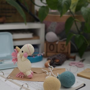 May include: A small cream-colored crocheted unicorn toy with a yellow horn, pink mane, and pink hooves, standing on a brown envelope next to two small crocheted spheres in yellow and teal, all resting on an open spiral-bound notebook.