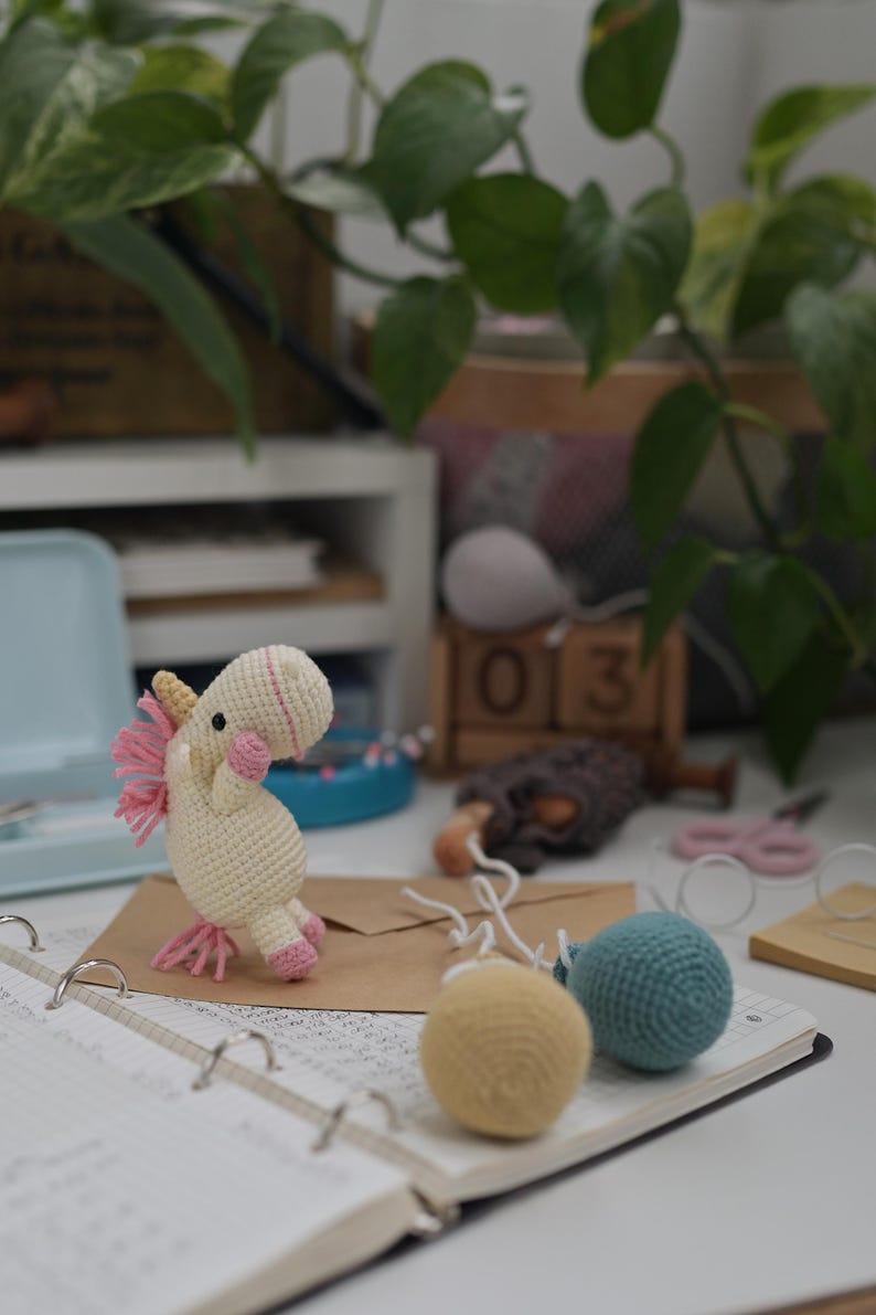 May include: A small cream-colored crocheted unicorn toy with a yellow horn, pink mane, and pink hooves, standing on a brown envelope next to two small crocheted spheres in yellow and teal, all resting on an open spiral-bound notebook.