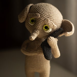 Puede incluir: Un juguete de peluche tejido a crochet con un cuerpo marr&oacute;n, ojos grandes verdes y una nariz larga gris. El juguete est&aacute; sosteniendo un objeto negro tejido a crochet en sus manos.