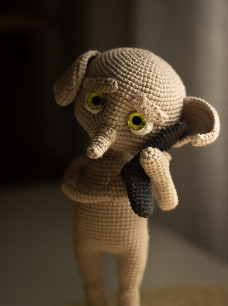 Puede incluir: Un juguete de peluche tejido a crochet con un cuerpo marr&oacute;n, ojos grandes verdes y una nariz larga gris. El juguete est&aacute; sosteniendo un objeto negro tejido a crochet en sus manos.