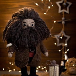Pode incluir: Um brinquedo de pel&uacute;cia de croch&ecirc; de um mago com uma longa barba marrom e cabelo marrom. O brinquedo est&aacute; vestindo um casaco marrom e uma camisa vermelha. O brinquedo est&aacute; em p&eacute; em uma superf&iacute;cie marrom com uma pequena lanterna de croch&ecirc;.