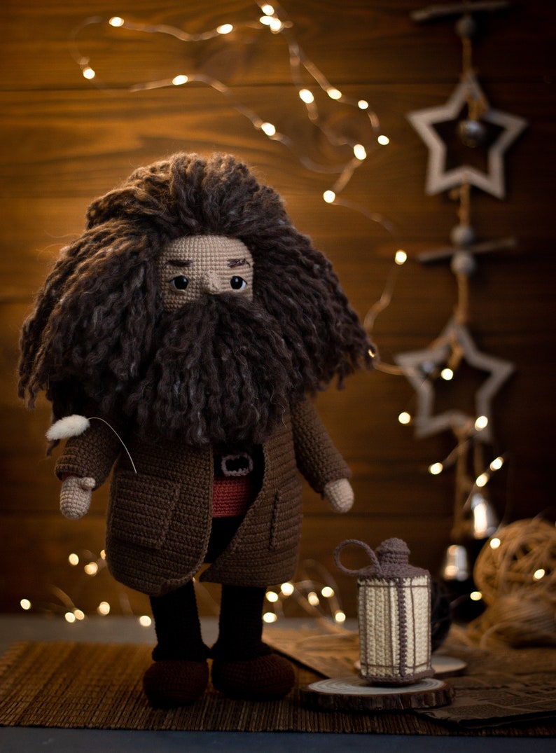 Pode incluir: Um brinquedo de pel&uacute;cia de croch&ecirc; de um mago com uma longa barba marrom e cabelo marrom. O brinquedo est&aacute; vestindo um casaco marrom e uma camisa vermelha. O brinquedo est&aacute; em p&eacute; em uma superf&iacute;cie marrom com uma pequena lanterna de croch&ecirc;.