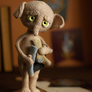 Puede incluir: Una mu&ntilde;eca de Dobby el elfo dom&eacute;stico de crochet con una expresi&oacute;n triste, sosteniendo un par de calcetines en sus manos. La mu&ntilde;eca es de color marr&oacute;n claro con ojos verdes y una nariz larga y puntiaguda.