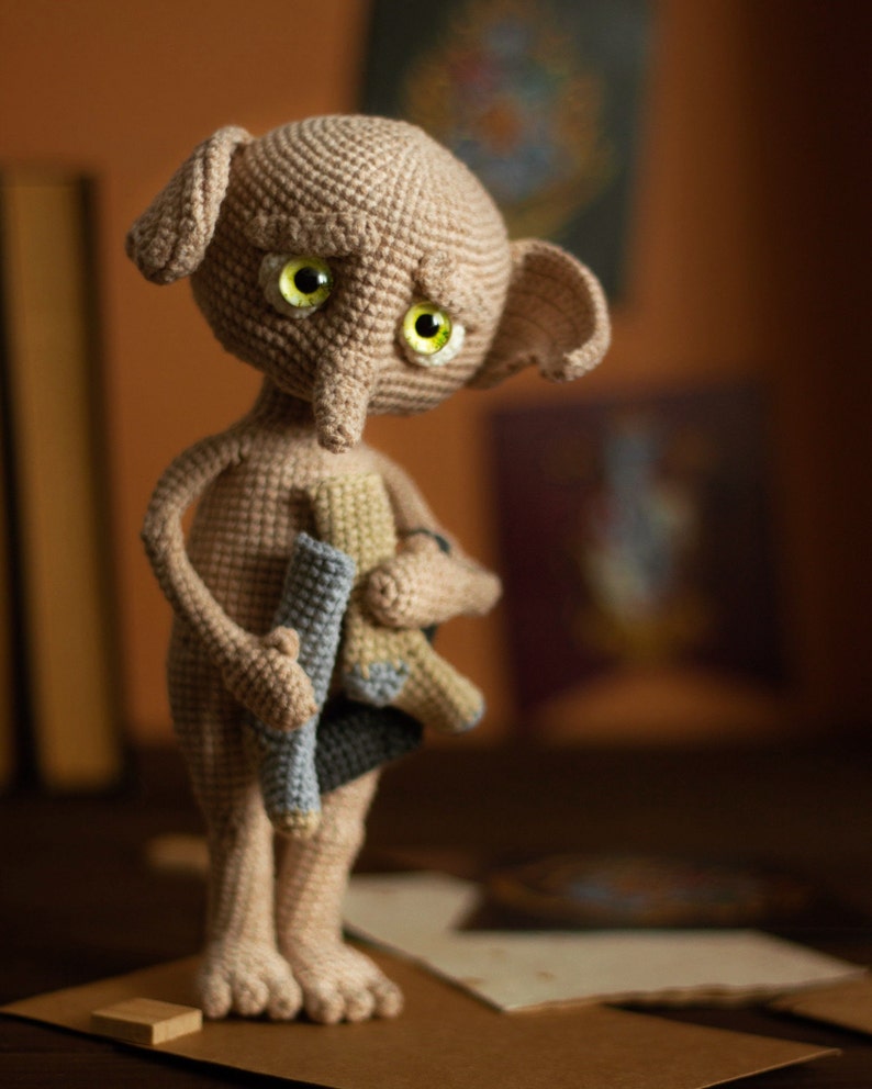 Puede incluir: Una mu&ntilde;eca de Dobby el elfo dom&eacute;stico de crochet con una expresi&oacute;n triste, sosteniendo un par de calcetines en sus manos. La mu&ntilde;eca es de color marr&oacute;n claro con ojos verdes y una nariz larga y puntiaguda.
