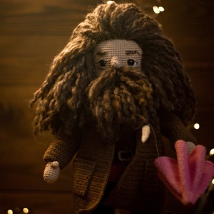 Pode incluir: Uma boneca de croch&ecirc; com cabelo castanho e barba, vestindo um casaco marrom e segurando um guarda-chuva rosa de croch&ecirc;. A boneca est&aacute; em p&eacute; em frente a um fundo de madeira com luzes.