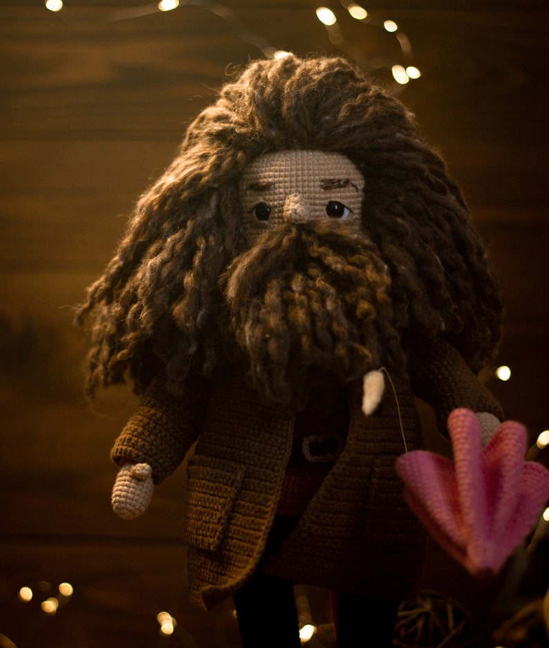 Pode incluir: Uma boneca de croch&ecirc; com cabelo castanho e barba, vestindo um casaco marrom e segurando um guarda-chuva rosa de croch&ecirc;. A boneca est&aacute; em p&eacute; em frente a um fundo de madeira com luzes.