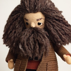 Pode incluir: Boneca amigurumi de croch&ecirc; de um personagem com cabelo castanho, barba castanha e casaco castanho. A boneca est&aacute; vestindo uma camisa vermelha e cal&ccedil;as pretas.