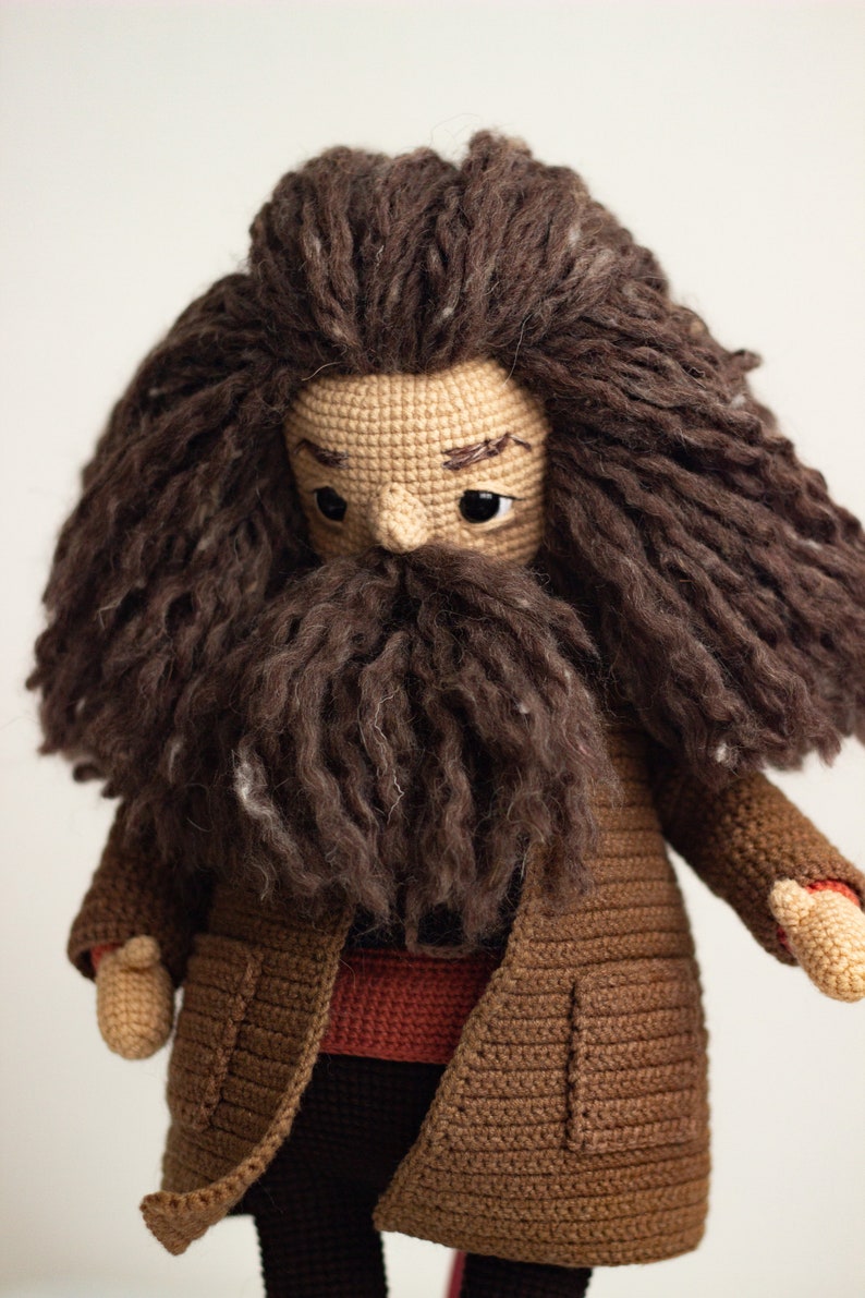 Pode incluir: Boneca amigurumi de croch&ecirc; de um personagem com cabelo castanho, barba castanha e casaco castanho. A boneca est&aacute; vestindo uma camisa vermelha e cal&ccedil;as pretas.