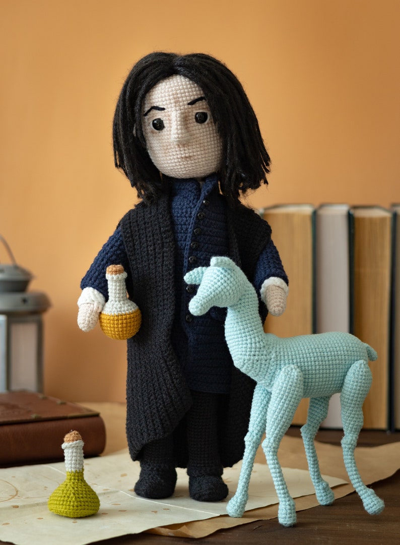 May include: Crocheted doll of a fictional character with long black hair, wearing a black coat and blue shirt, holding a yellow potion bottle. A blue crocheted animal stands next to the doll.