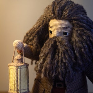 Pode incluir: Um brinquedo de pel&uacute;cia de croch&ecirc; de um mago com uma longa barba e cabelo castanho, segurando uma lanterna. O brinquedo est&aacute; vestindo um casaco marrom e tem uma express&atilde;o amig&aacute;vel.