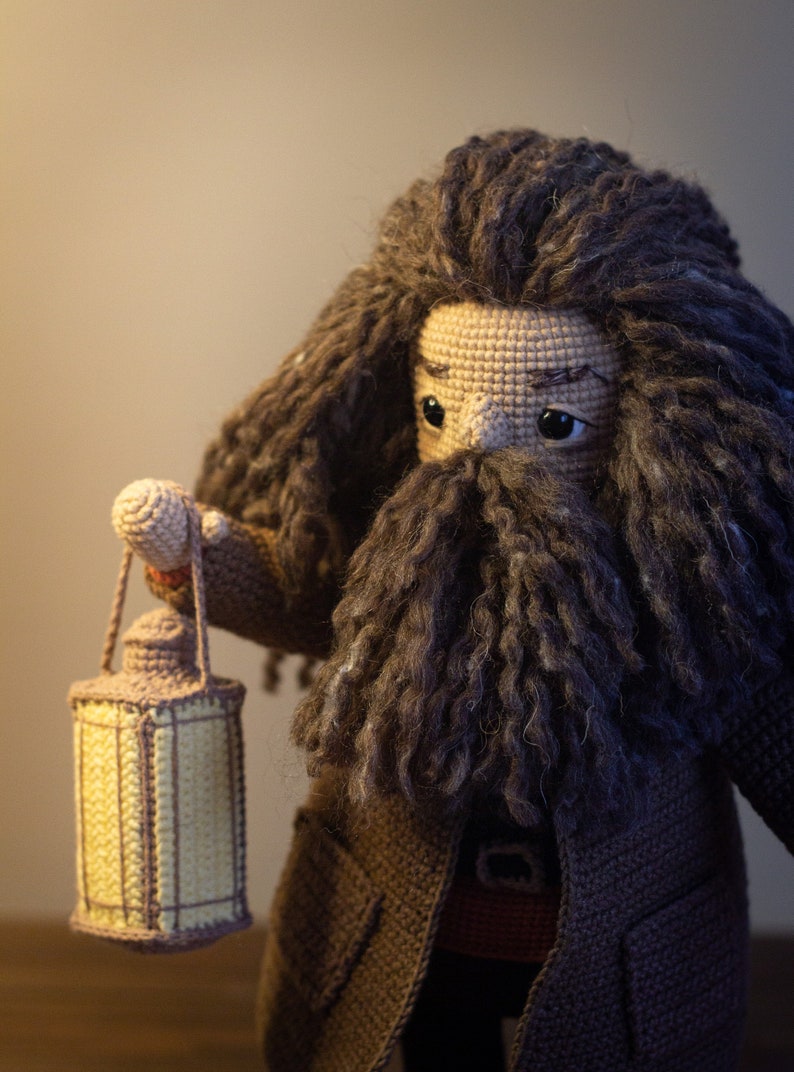 Pode incluir: Um brinquedo de pel&uacute;cia de croch&ecirc; de um mago com uma longa barba e cabelo castanho, segurando uma lanterna. O brinquedo est&aacute; vestindo um casaco marrom e tem uma express&atilde;o amig&aacute;vel.