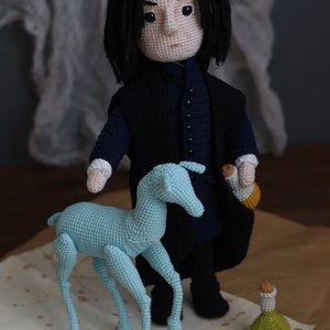 May include: A crocheted doll with black hair and a black robe, standing next to a light blue crocheted animal. The doll is holding a small crocheted bottle. The doll and animal are on a brown surface with white splotches.