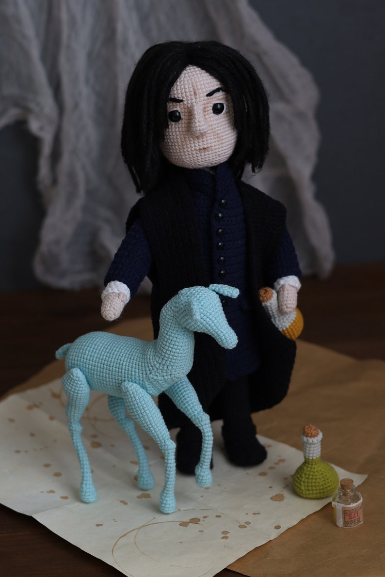 May include: A crocheted doll with black hair and a black robe, standing next to a light blue crocheted animal. The doll is holding a small crocheted bottle. The doll and animal are on a brown surface with white splotches.
