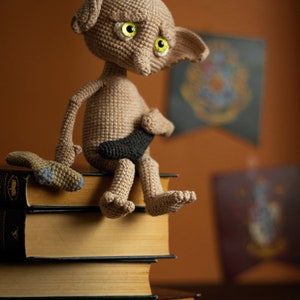 Puede incluir: Una mu&ntilde;eca de Dobby el elfo dom&eacute;stico de crochet sentada en una pila de libros. La mu&ntilde;eca tiene grandes ojos verdes y un cuerpo marr&oacute;n. La mu&ntilde;eca sostiene una varita negra en su mano.