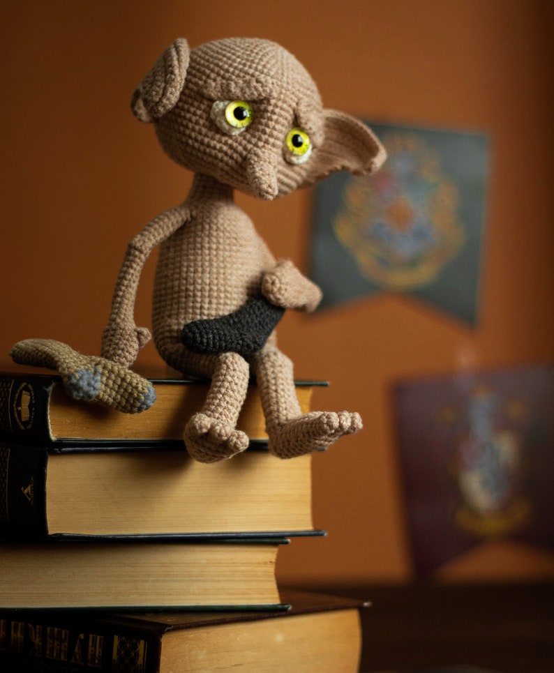 Puede incluir: Una mu&ntilde;eca de Dobby el elfo dom&eacute;stico de crochet sentada en una pila de libros. La mu&ntilde;eca tiene grandes ojos verdes y un cuerpo marr&oacute;n. La mu&ntilde;eca sostiene una varita negra en su mano.