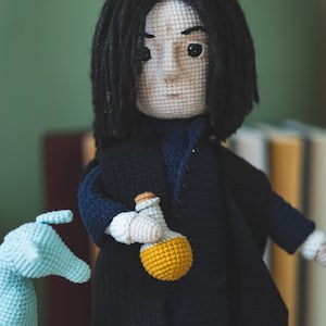 May include: Crocheted doll with long black hair wearing a dark blue coat and holding a yellow bottle. The doll is standing on a white surface.