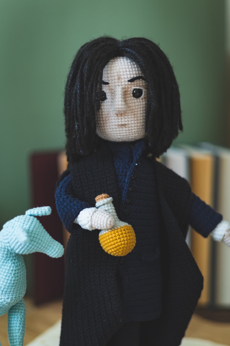 May include: Crocheted doll with long black hair wearing a dark blue coat and holding a yellow bottle. The doll is standing on a white surface.