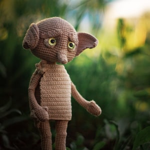 Puede incluir: Un juguete de peluche de crochet de un elfo dom&eacute;stico con orejas grandes, nariz puntiaguda y grandes ojos verdes. El elfo est&aacute; de pie sobre una cama de hojas verdes.