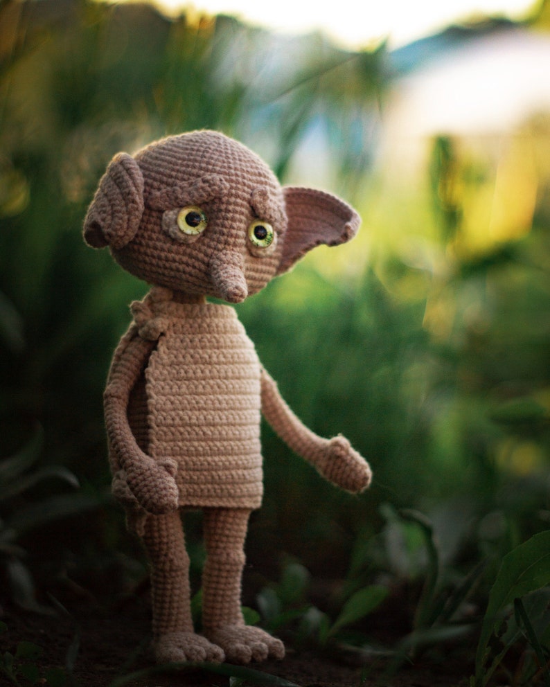 Puede incluir: Un juguete de peluche de crochet de un elfo dom&eacute;stico con orejas grandes, nariz puntiaguda y grandes ojos verdes. El elfo est&aacute; de pie sobre una cama de hojas verdes.