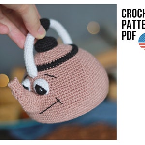 Könnte beinhalten: Eine gehäkelte rosa Teekanne mit schwarzen und weißen Details. Die Teekanne hat ein lächelndes Gesicht mit großen Augen und einem kleinen Ausguss. Die Teekanne wird von einer Hand gehalten.