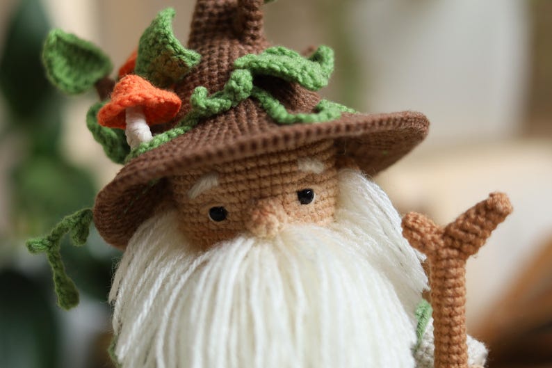 May include: A crocheted gnome with a brown hat, white beard, and a green leafy crown. The gnome is holding a brown stick in its right hand.