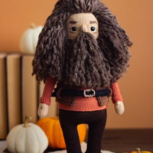 Pode incluir: Um brinquedo de pel&uacute;cia de croch&ecirc; de um personagem com uma longa barba e cabelo castanho, vestindo uma camisa vermelha, um colete marrom e cal&ccedil;as marrons. O brinquedo est&aacute; em p&eacute; sobre uma superf&iacute;cie de madeira com ab&oacute;boras.