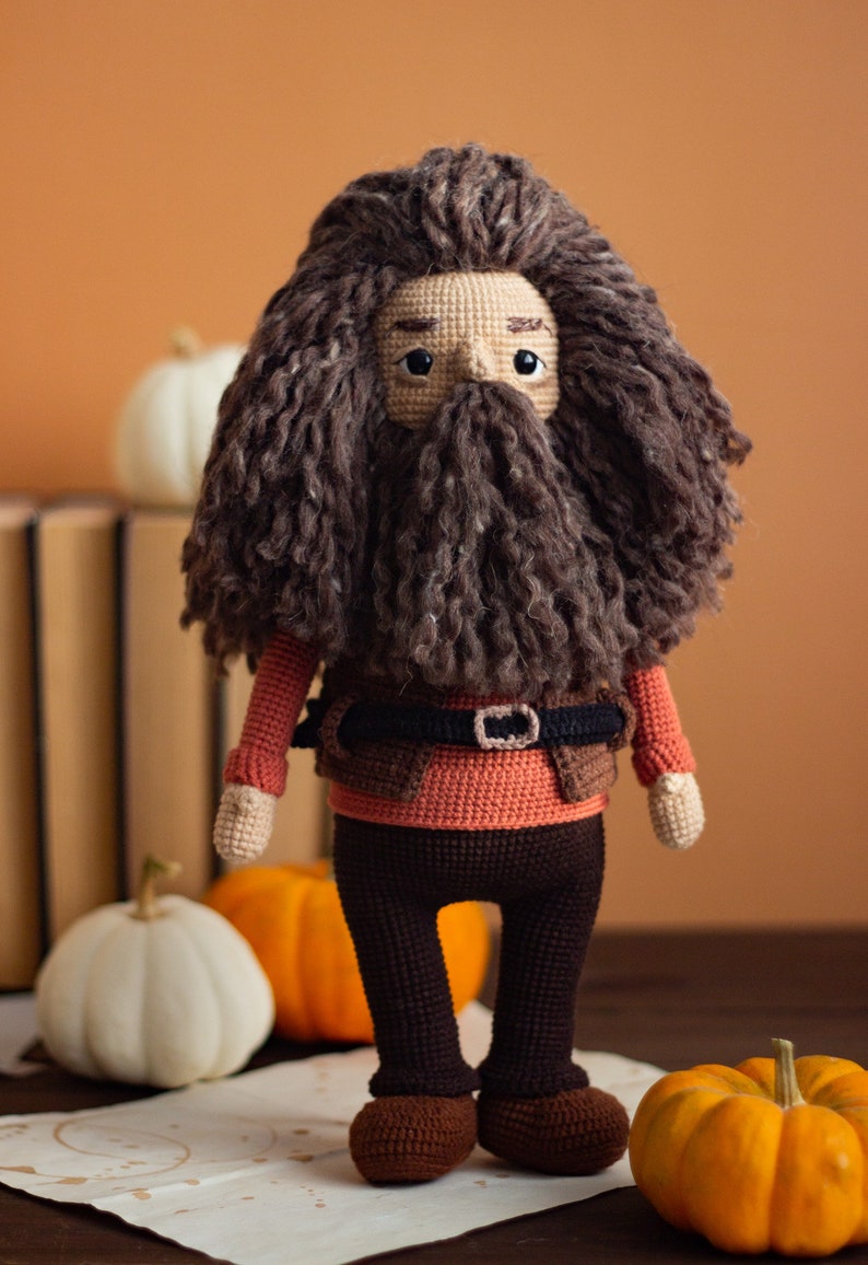 Pode incluir: Um brinquedo de pel&uacute;cia de croch&ecirc; de um personagem com uma longa barba e cabelo castanho, vestindo uma camisa vermelha, um colete marrom e cal&ccedil;as marrons. O brinquedo est&aacute; em p&eacute; sobre uma superf&iacute;cie de madeira com ab&oacute;boras.