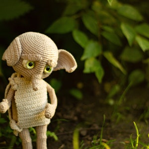 Puede incluir: Un juguete de peluche de crochet de un elfo dom&eacute;stico con grandes ojos verdes y una nariz puntiaguda. El elfo est&aacute; de pie sobre una cama de hierba verde.