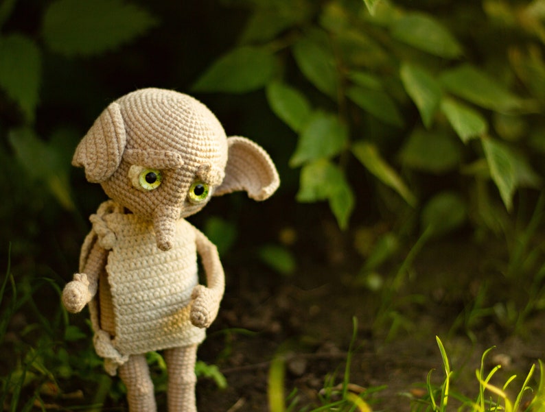 Puede incluir: Un juguete de peluche de crochet de un elfo dom&eacute;stico con grandes ojos verdes y una nariz puntiaguda. El elfo est&aacute; de pie sobre una cama de hierba verde.