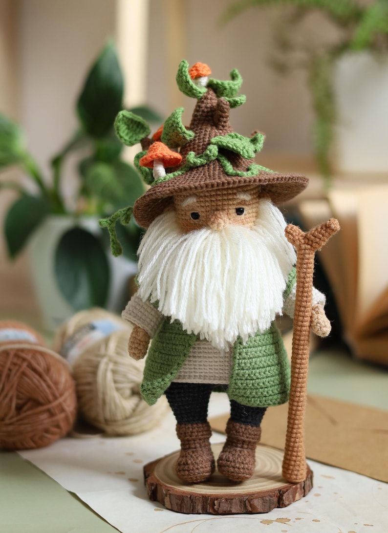 May include: A crocheted gnome with a brown hat and a white beard, wearing a green jacket and brown boots. The gnome is holding a brown stick and is standing on a wooden base. The gnome's hat is decorated with green leaves and orange mushrooms.