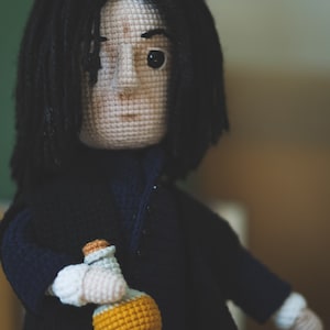 May include: Crocheted doll with black hair, wearing a dark blue coat and holding a yellow potion bottle.