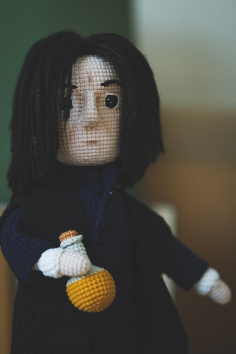 May include: Crocheted doll with black hair, wearing a dark blue coat and holding a yellow potion bottle.