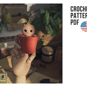 Puede incluir: Una planta de Mandrágora tejida a ganchillo en una maceta roja, sostenida por una mano de madera. La Mandrágora tiene una cara sonriente y hojas verdes. La imagen incluye el texto "CROCHET PATTERN PDF" y una bandera americana.