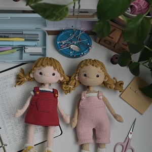 May include: Two handmade crochet dolls with blonde yarn hair styled in pigtails. One doll wears a red overall dress with yellow sneakers, while the other wears pink overalls and white socks. Both dolls feature embroidered facial details and light skin.