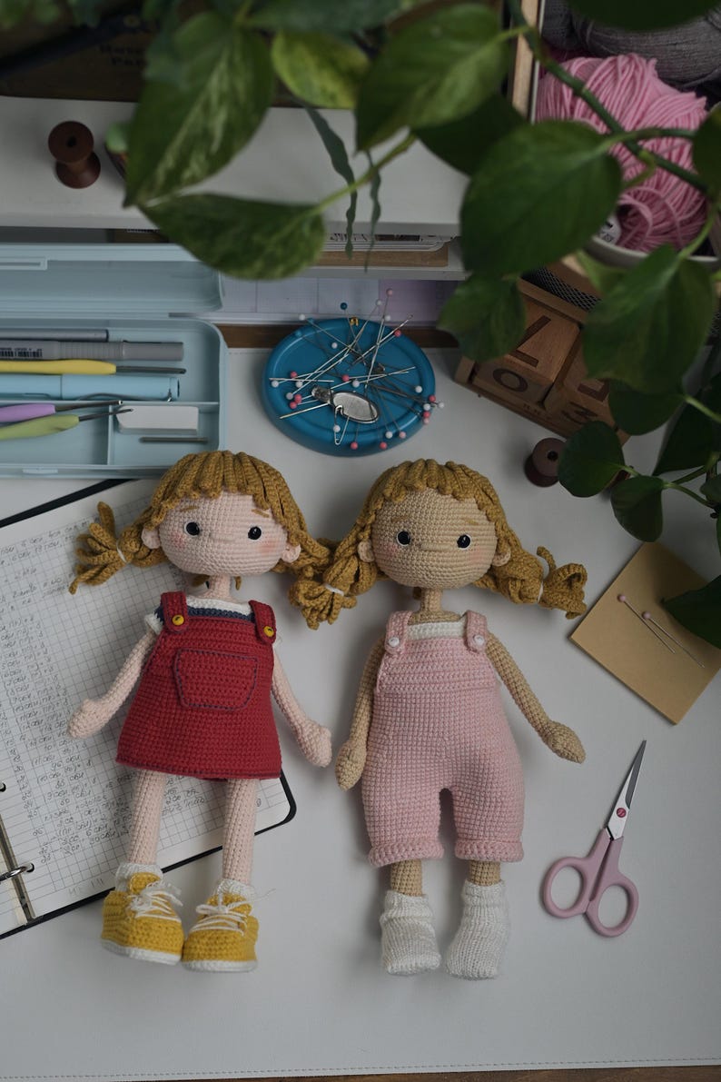 May include: Two handmade crochet dolls with blonde yarn hair styled in pigtails. One doll wears a red overall dress with yellow sneakers, while the other wears pink overalls and white socks. Both dolls feature embroidered facial details and light skin.