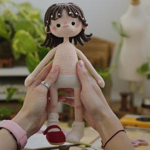 May include: A handmade crochet doll with dark brown hair, small white beads, and rosy cheeks. The doll has a light beige skin tone, wears a light-colored diaper, one white sock, and one red Mary Jane shoe. The doll features black bead eyes.
