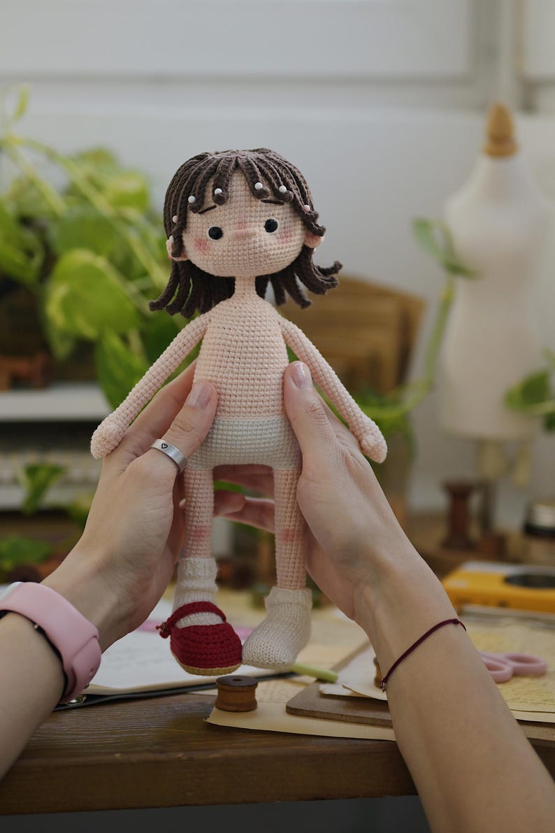 May include: A handmade crochet doll with dark brown hair, small white beads, and rosy cheeks. The doll has a light beige skin tone, wears a light-colored diaper, one white sock, and one red Mary Jane shoe. The doll features black bead eyes.
