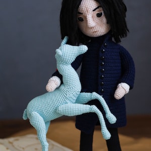 May include: A crocheted doll with long black hair wearing a dark blue coat and black pants. The doll is holding a light blue crocheted animal with four legs.