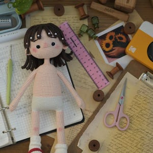 May include: A handmade crochet doll with dark brown hair, pink cheeks, and a light skin tone. The doll wears white underwear, one white sock, and one red shoe. A pink 6-inch ruler is positioned next to the doll, showing its scale against craft supplies.