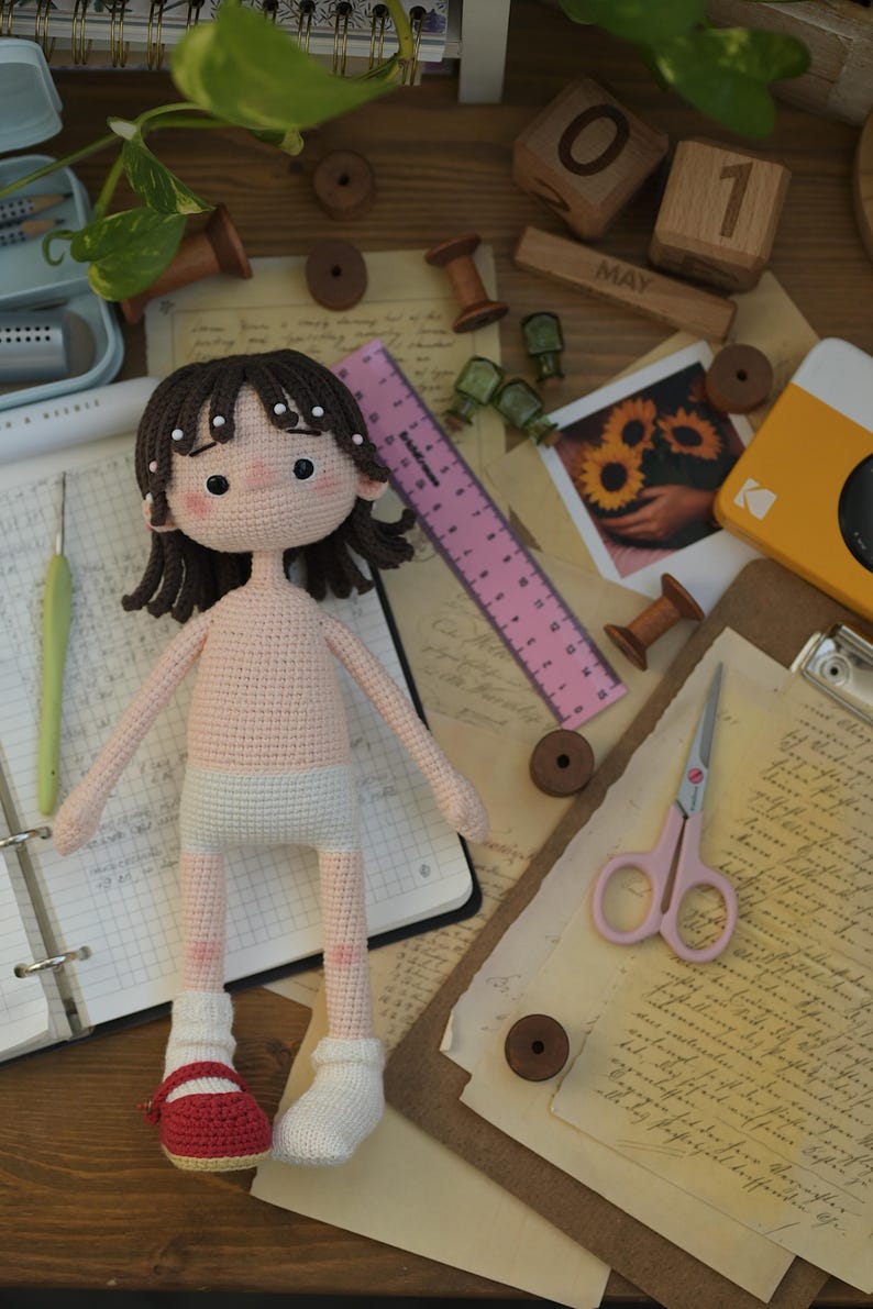 May include: A handmade crochet doll with dark brown hair, pink cheeks, and a light skin tone. The doll wears white underwear, one white sock, and one red shoe. A pink 6-inch ruler is positioned next to the doll, showing its scale against craft supplies.