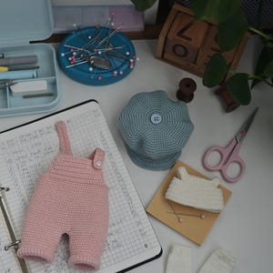 May include: A set of miniature crochet clothing items including a light pink pair of overalls with a single button, a dusty blue beret with a central button, a small white top, and two white leg warmers, arranged alongside sewing supplies.