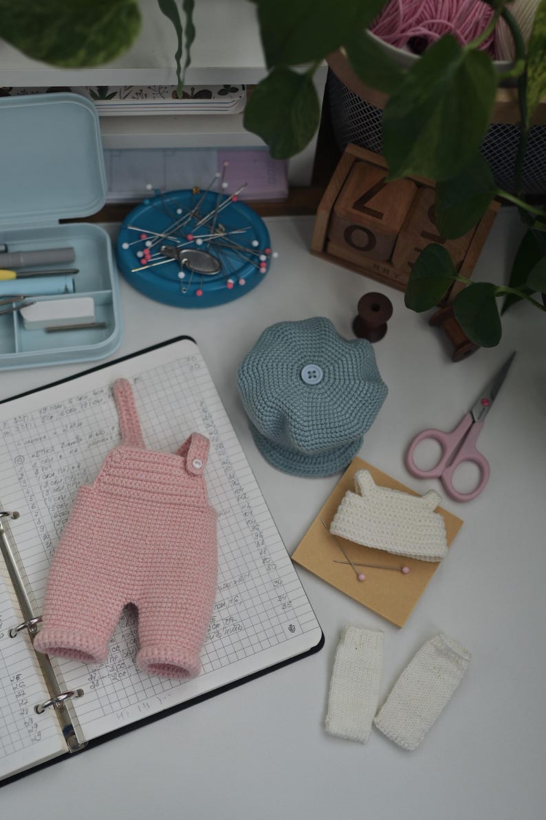 May include: A set of miniature crochet clothing items including a light pink pair of overalls with a single button, a dusty blue beret with a central button, a small white top, and two white leg warmers, arranged alongside sewing supplies.