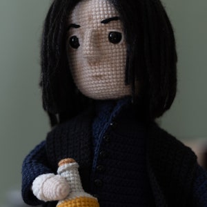 May include: Crocheted doll with long black hair, wearing a dark blue sweater and holding a small yellow crocheted bottle.