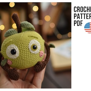 Può includere: Schema a uncinetto PDF per un giocattolo amigurumi a forma di mela verde con gambo marrone, braccia marroni e un viso carino con occhi bianchi e guance rosa.