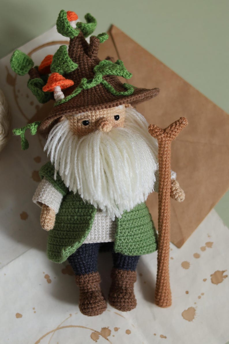 May include: A crocheted gnome with a long white beard, wearing a brown hat with green vines and orange mushrooms, holding a brown stick.