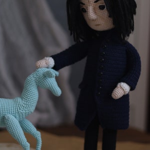 May include: A crocheted doll with long black hair wearing a dark blue coat and black pants. The doll is holding a light blue crocheted animal.
