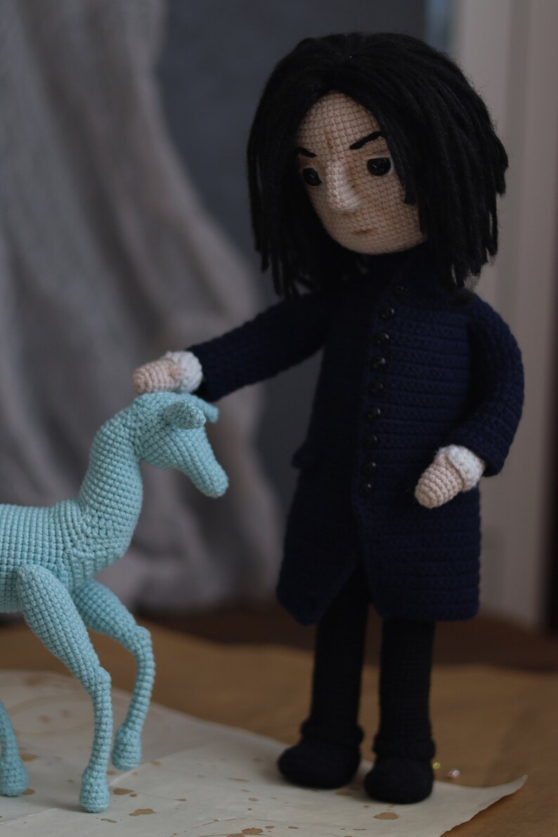 May include: A crocheted doll with long black hair wearing a dark blue coat and black pants. The doll is holding a light blue crocheted animal.